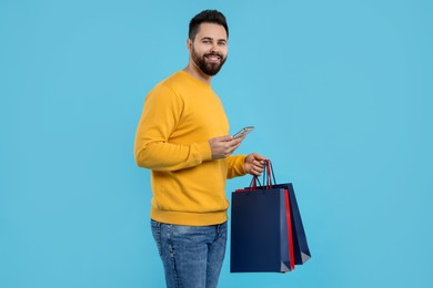 Smiling man with paper shopping bags and smartphone on light blue background