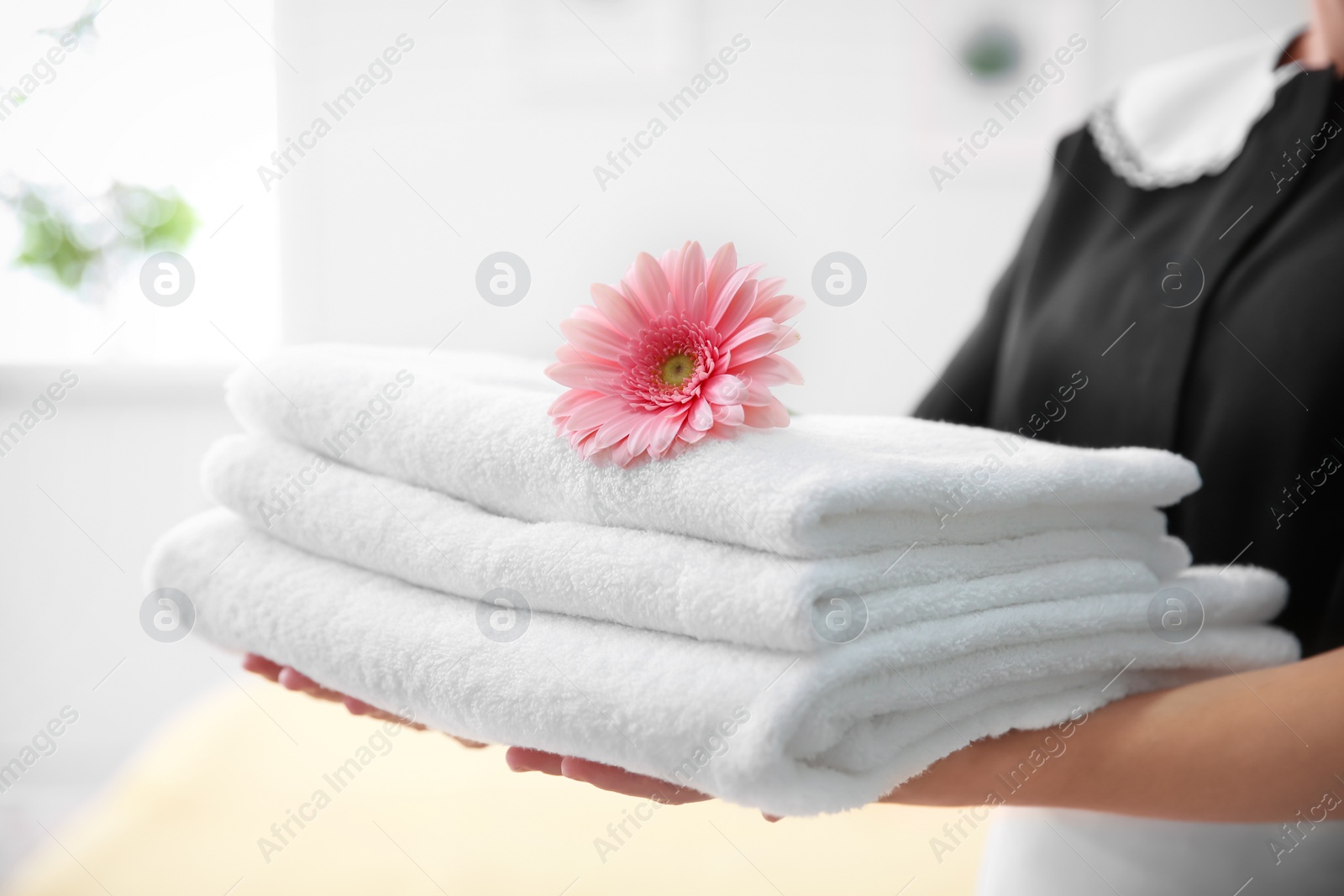 Photo of Young maid holding stack of towels and flower in hotel room