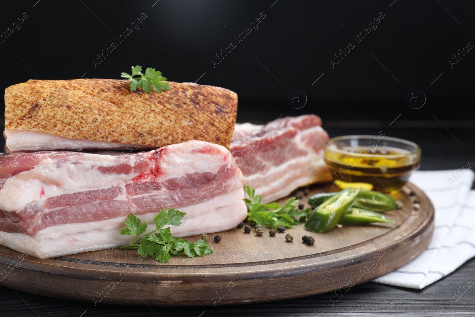Photo of Pieces of raw pork belly, chili pepper, peppercorns, oil and parsley on black wooden table, closeup