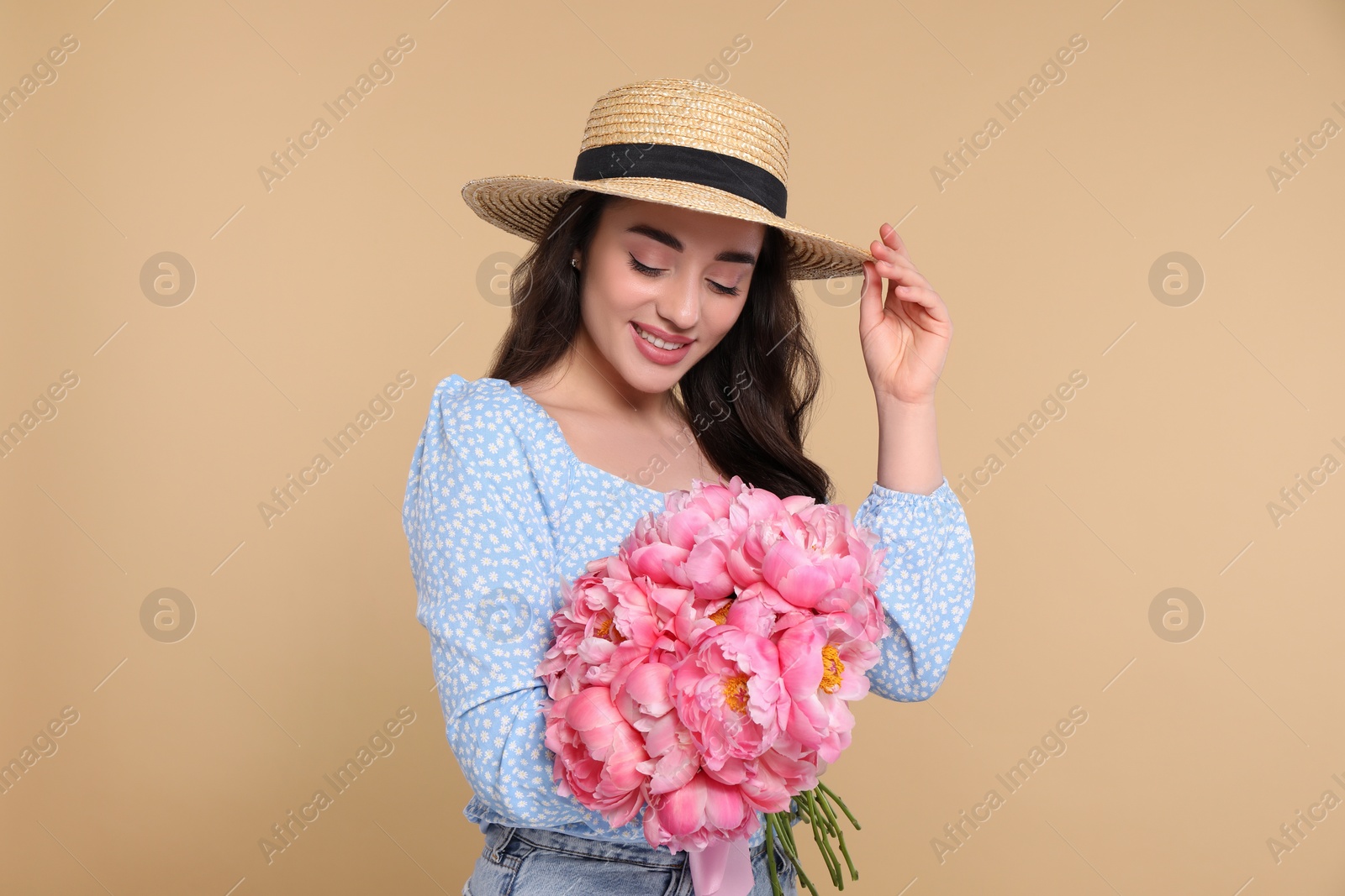 Photo of Beautiful young woman in straw hat with bouquet of pink peonies against beige background