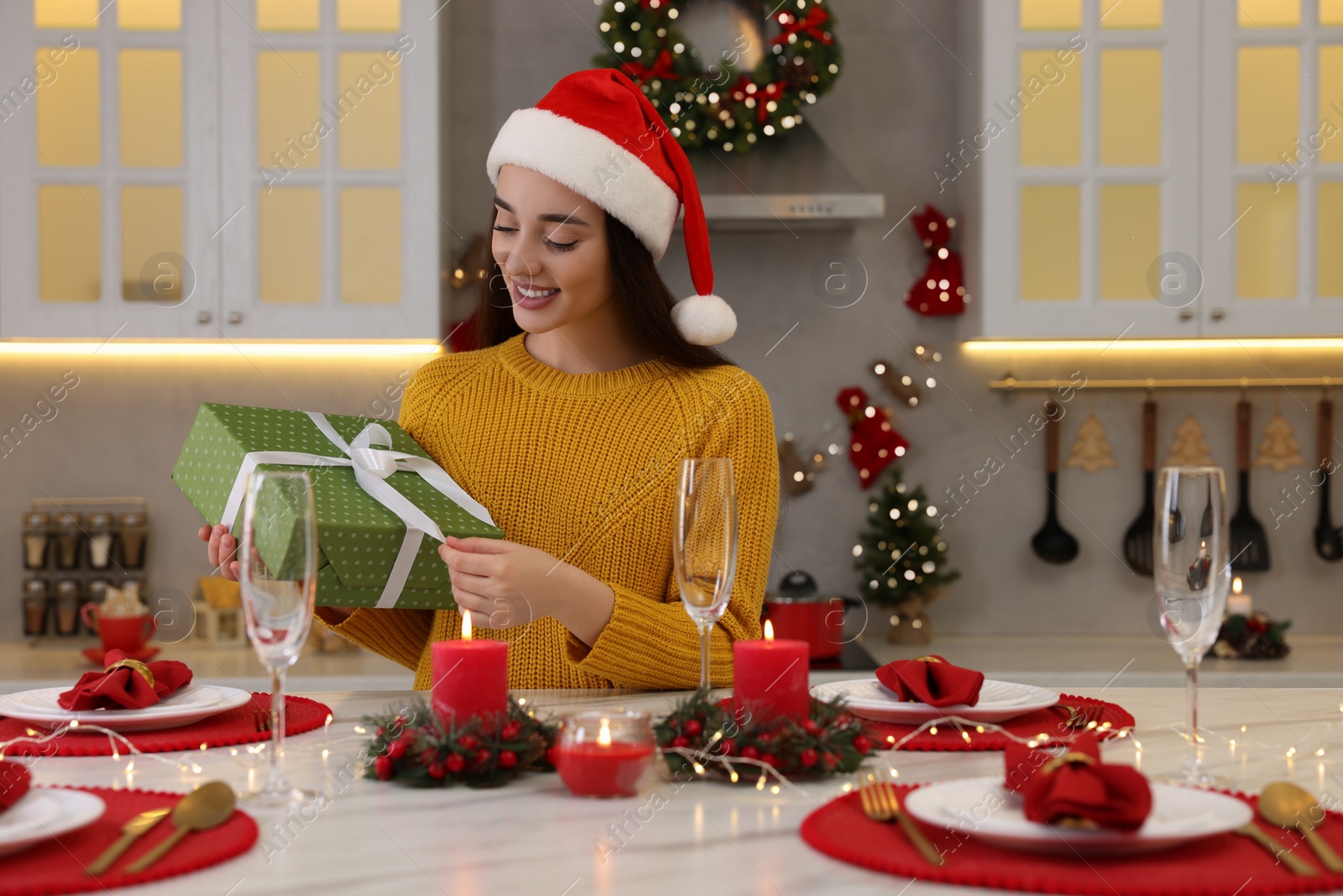 Photo of Happy young woman in Santa hat with Christmas gift at table in kitchen