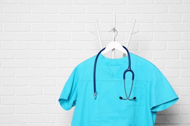 Turquoise medical uniform and stethoscope hanging on rack near white brick wall. Space for text