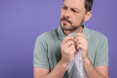 Angry man popping bubble wrap on purple background, closeup. Space for text. Stress relief