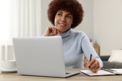 Photo of Beautiful young woman using laptop and writing in notebook at wooden desk in room