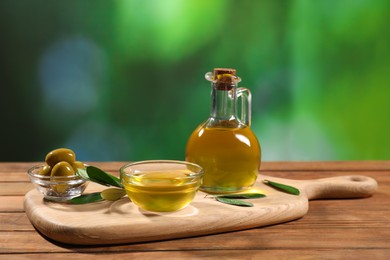 Photo of Cooking oil, olives and green leaves on wooden table against blurred background