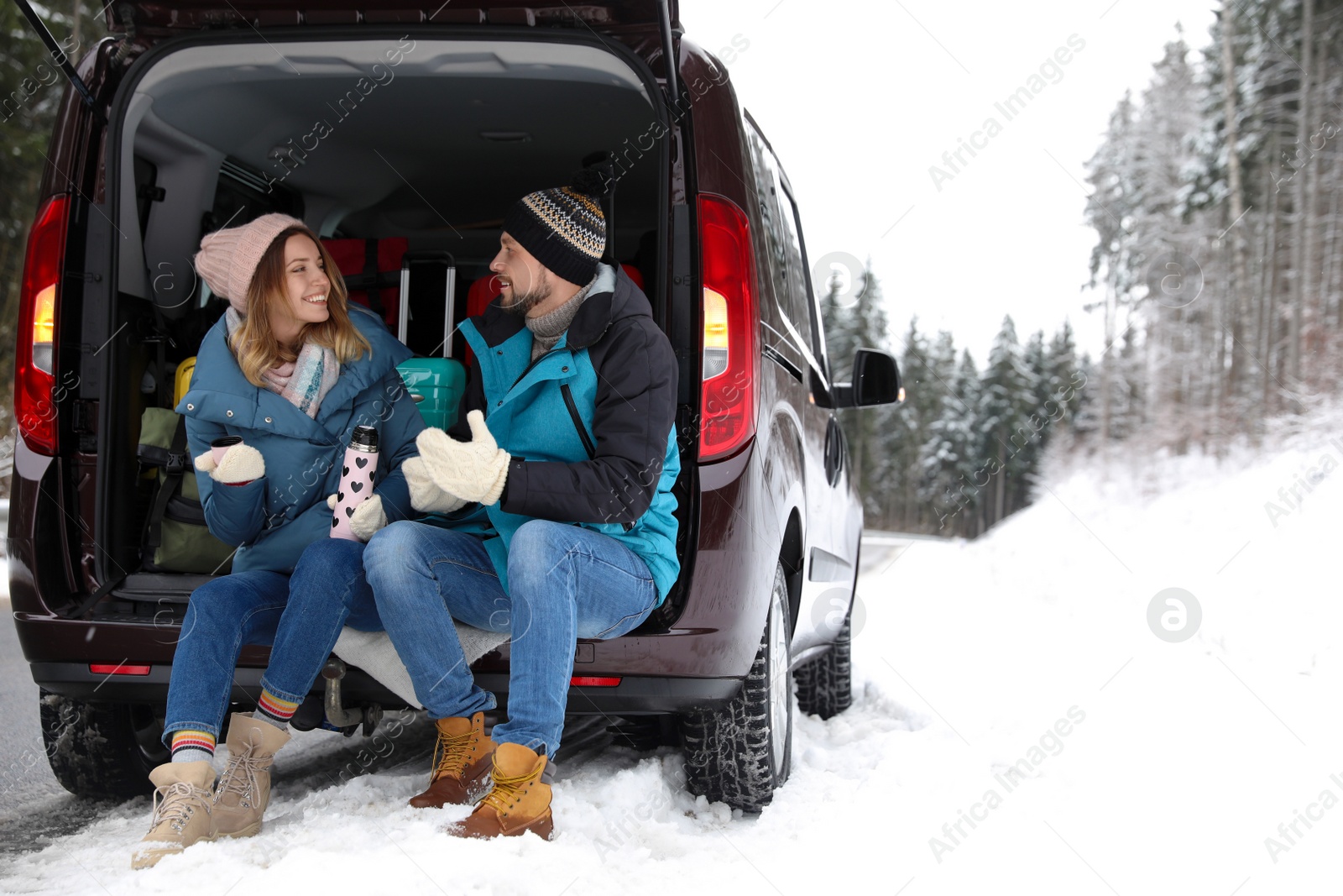 Photo of Couple sitting in open car trunk full of luggage near road, space for text. Winter vacation