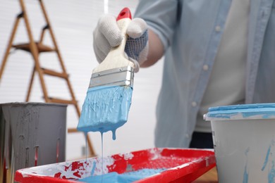 Photo of Man taking light blue paint with brush from tray indoors, closeup