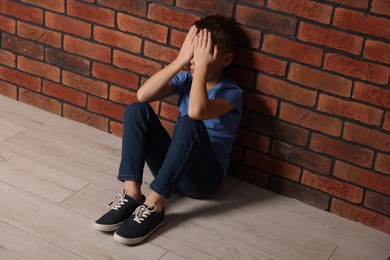 Photo of Child abuse. Upset boy sitting on floor near brick wall indoors