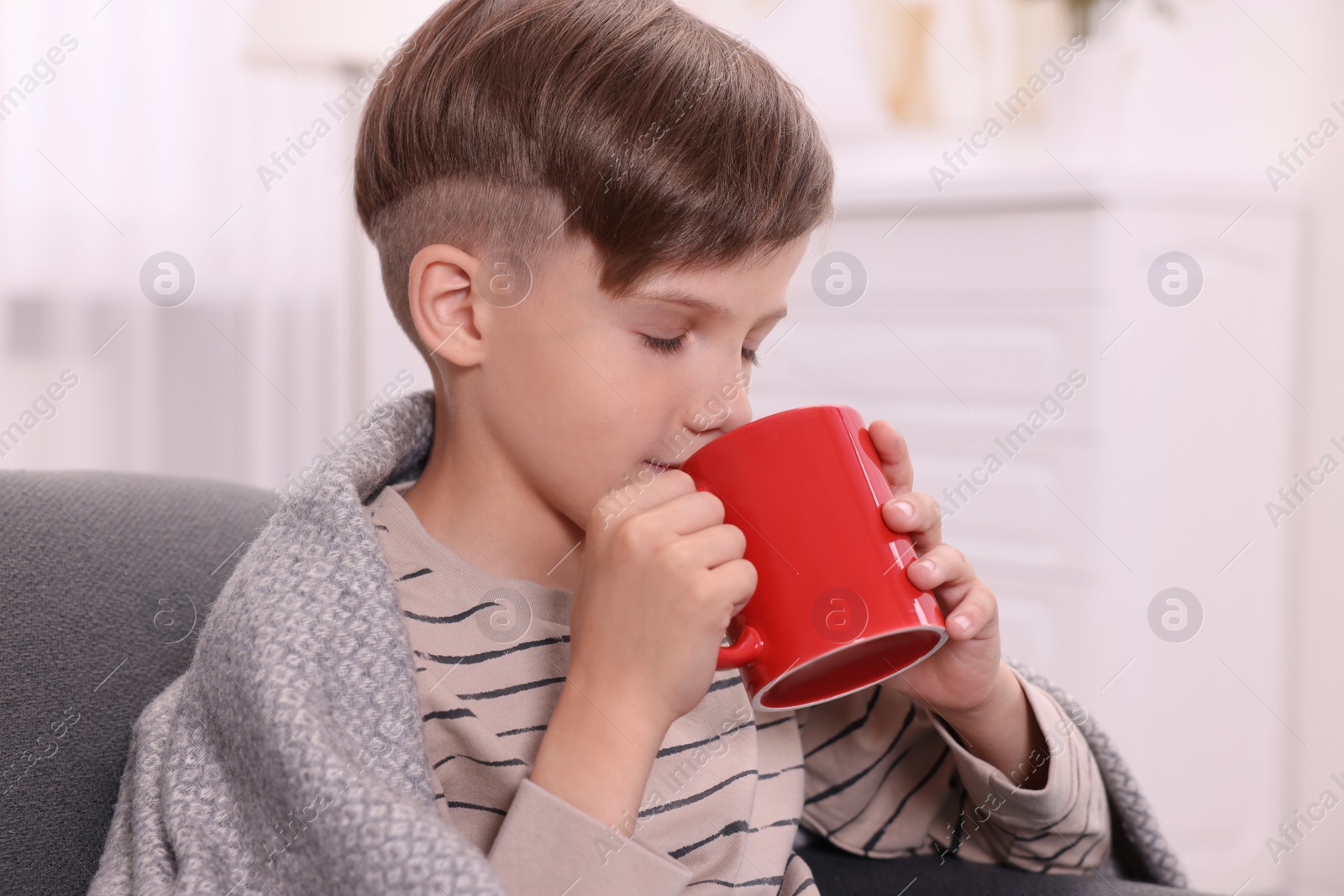 Photo of Cute boy drinking from red ceramic mug on sofa at home