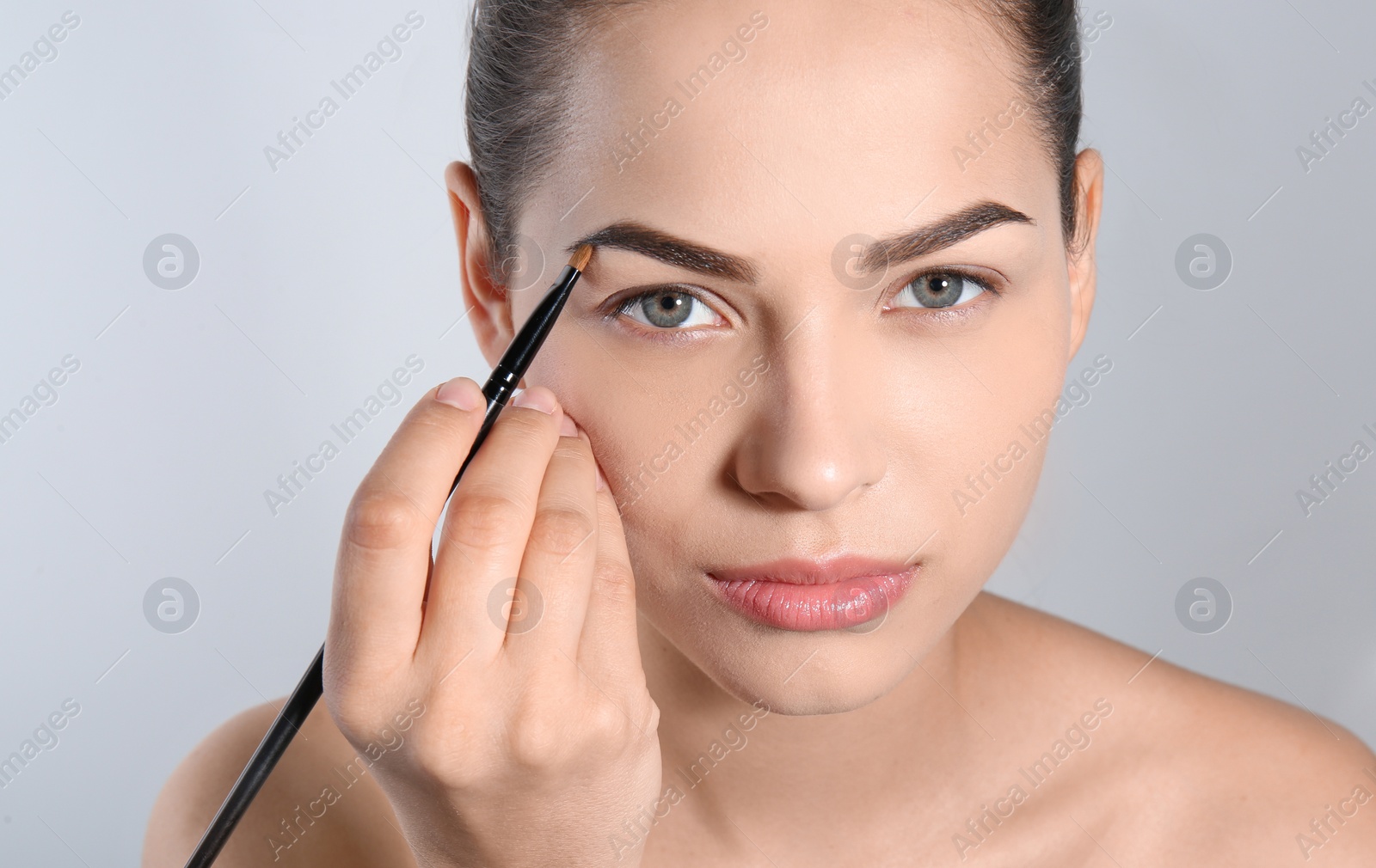 Photo of Young woman correcting shape of eyebrow with brush on light background