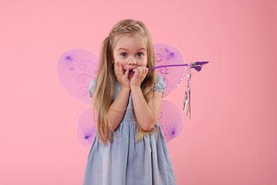 Surprised little girl in fairy costume with violet wings and magic wand on pink background