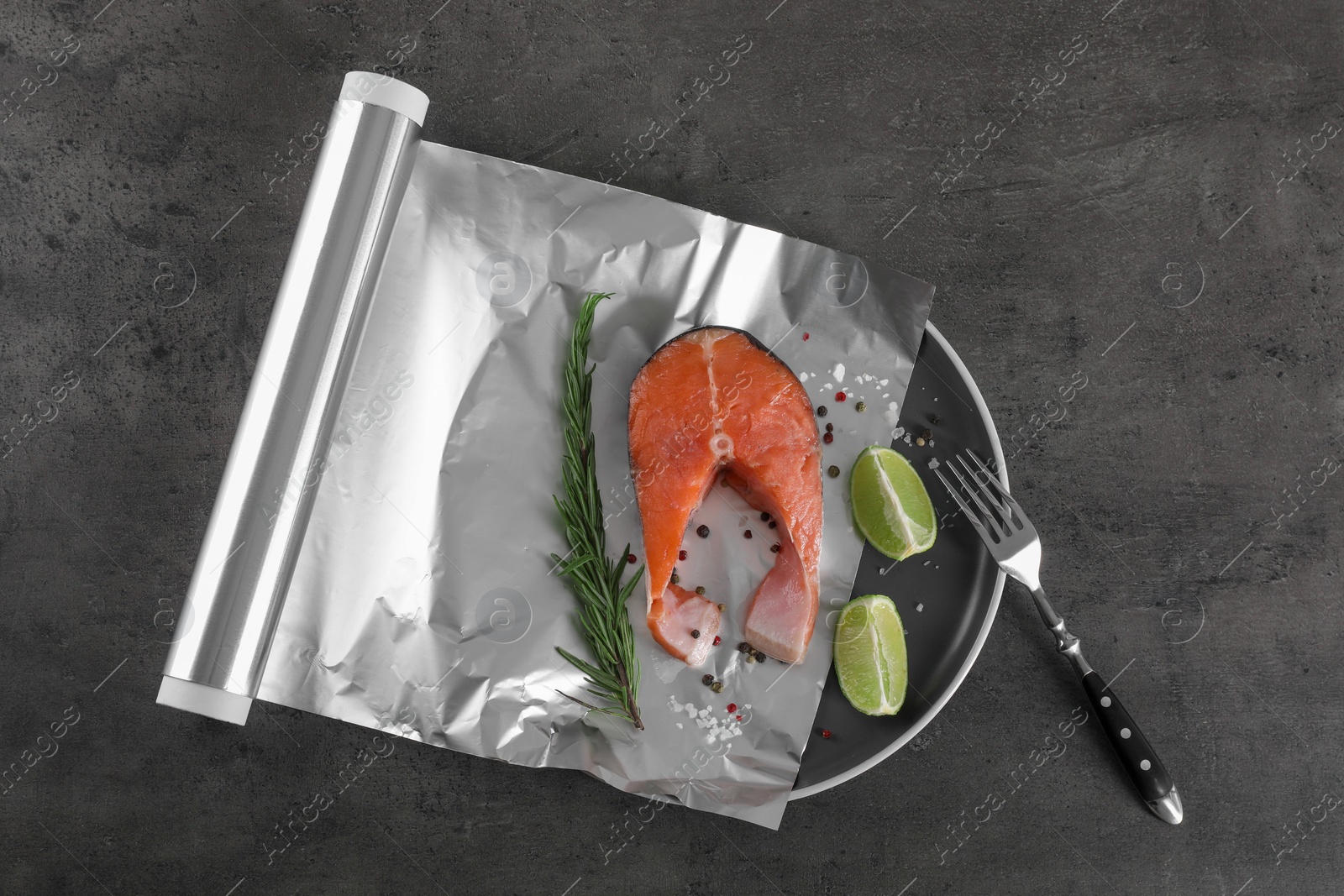 Photo of Aluminum foil with raw fish, lime, rosemary and spices on grey textured table, flat lay. Baking salmon