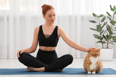 Beautiful woman petting cute red cat on mat at home. Practicing yoga