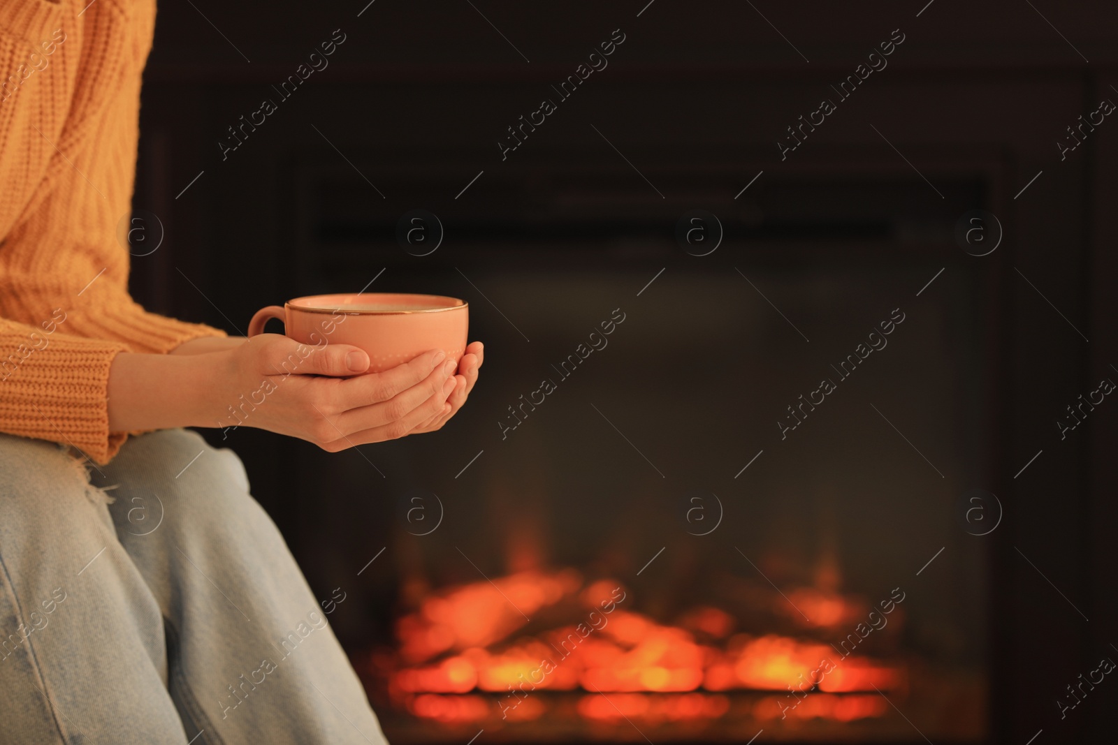 Photo of Young woman with cup of hot drink near fireplace, closeup. Cozy atmosphere