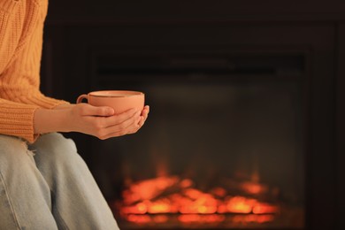 Young woman with cup of hot drink near fireplace, closeup. Cozy atmosphere