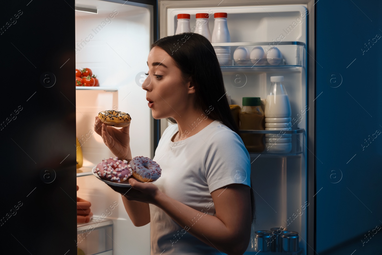 Photo of Young woman eating donut near refrigerator in kitchen at night. Bad habit