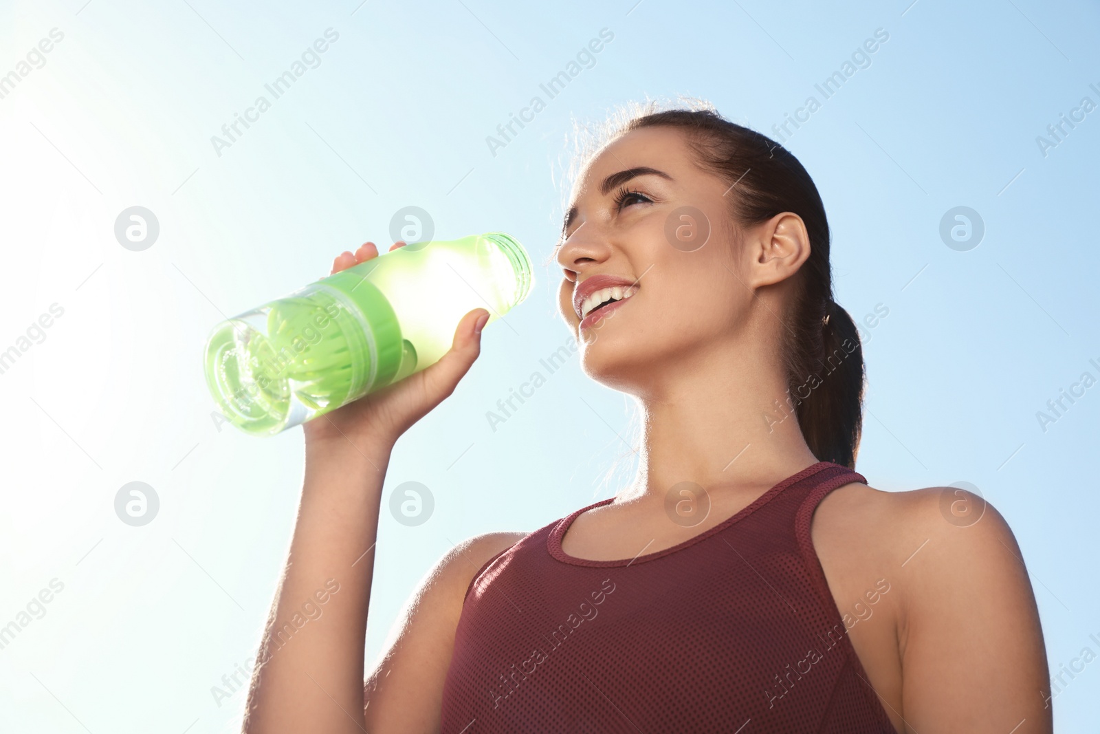 Photo of Young sporty woman drinking water from bottle against blue sky on sunny day