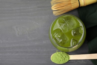 Photo of Glass of delicious iced green matcha tea, spoon with powder and bamboo whisk on grey wooden table, flat lay. Space for text