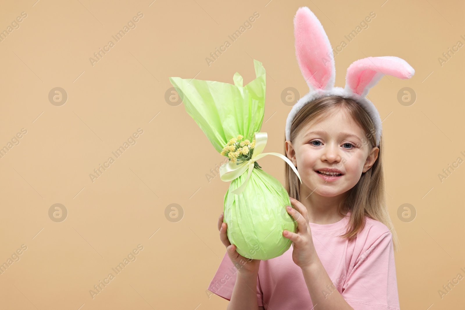 Photo of Easter celebration. Cute girl with bunny ears holding wrapped gift on beige background, space for text