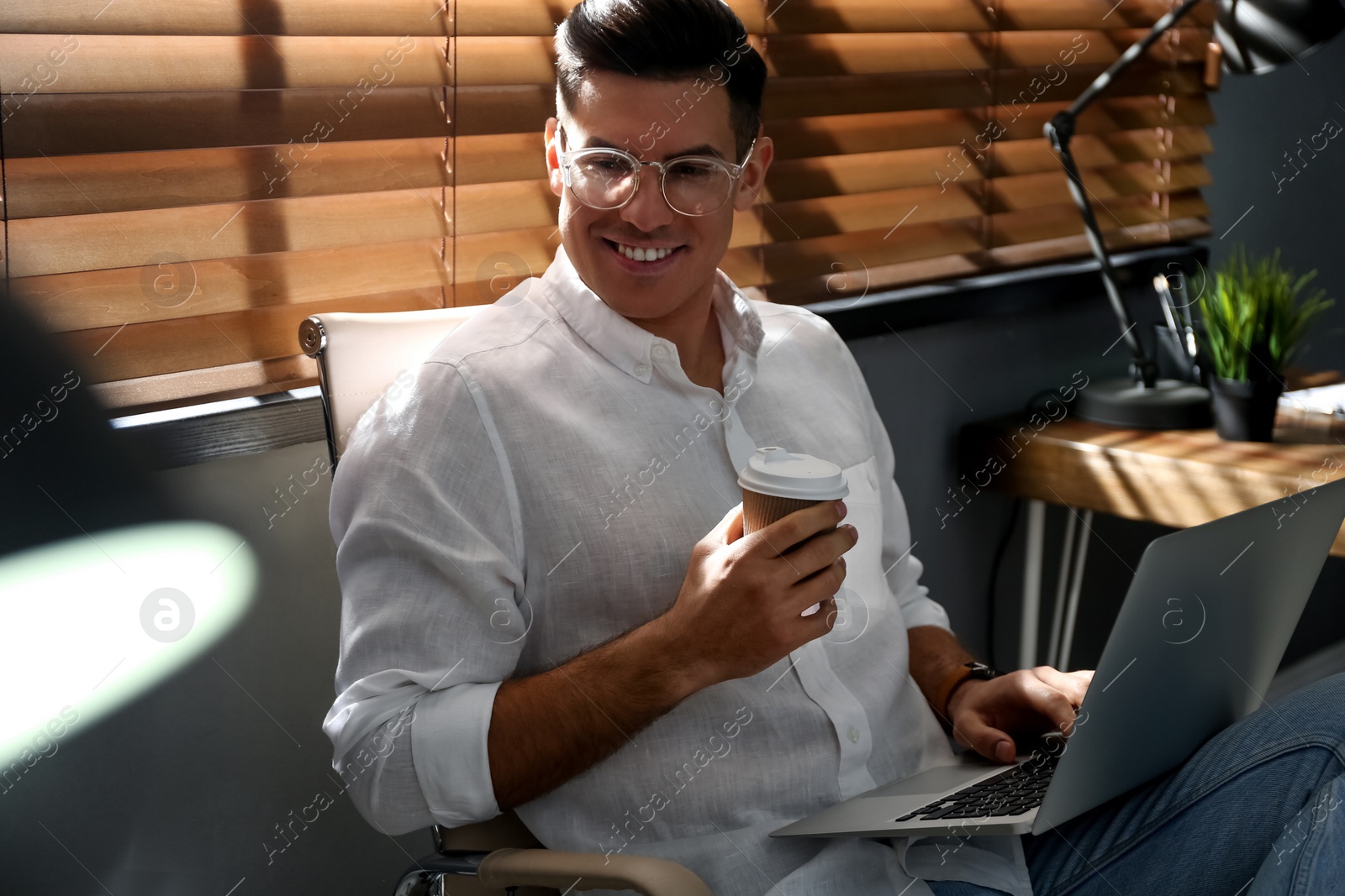 Photo of Freelancer with cup of coffee working on laptop near window indoors