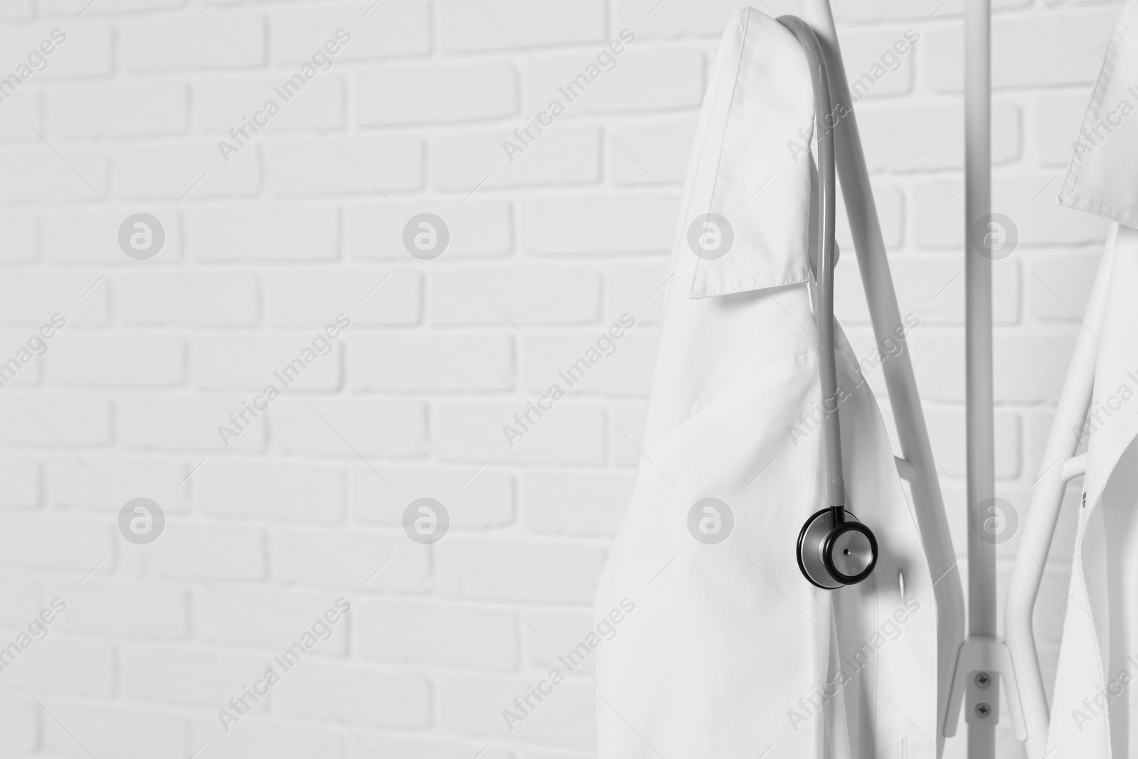 Photo of Medical uniforms and stethoscope hanging on rack near white brick wall. Space for text