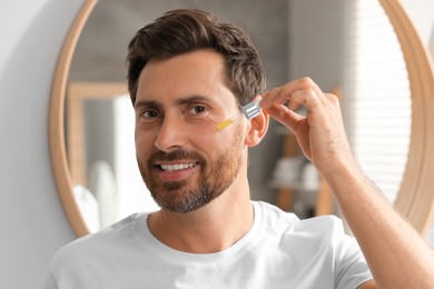 Smiling man applying cosmetic serum onto his face indoors. Space for text