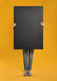 Woman holding black blank poster on yellow background. Mockup for design