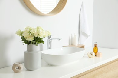 Vase with beautiful white roses and toiletries near sink in bathroom