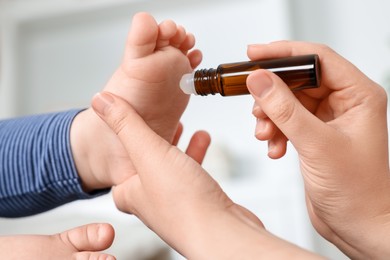 Photo of Mother applying essential oil from roller bottle onto her baby`s heel on blurred background, closeup