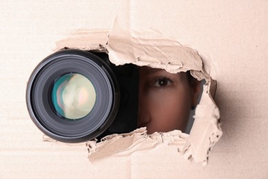 Photo of Hidden woman with camera spying through torn hole in cardboard, closeup