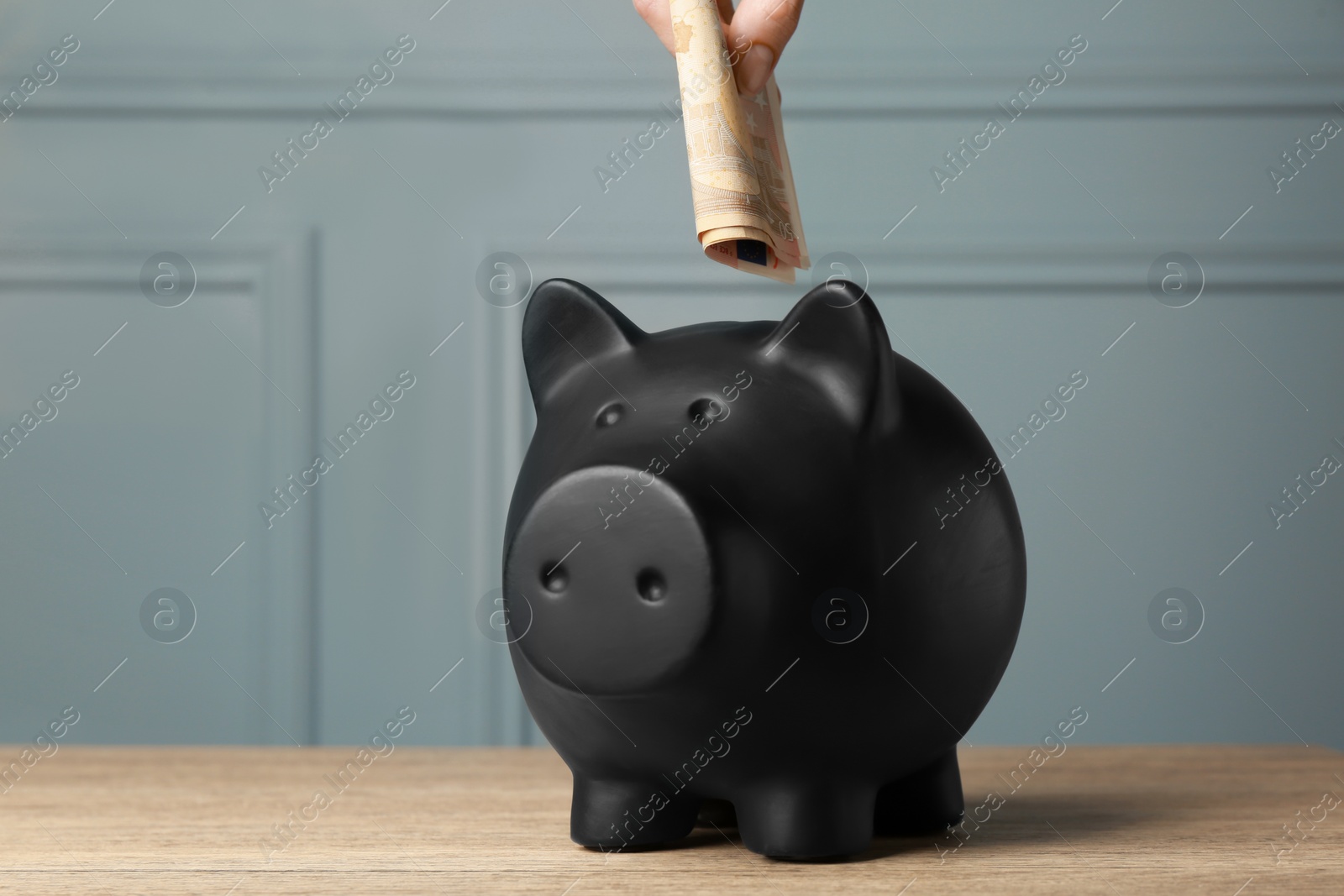 Photo of Woman putting banknotes into piggy bank at wooden table, closeup