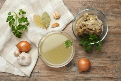 Photo of Glass bowl with delicious bone broth and ingredients on wooden table, flat lay