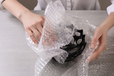 Woman covering ceramic bowl with bubble wrap at light grey table, closeup