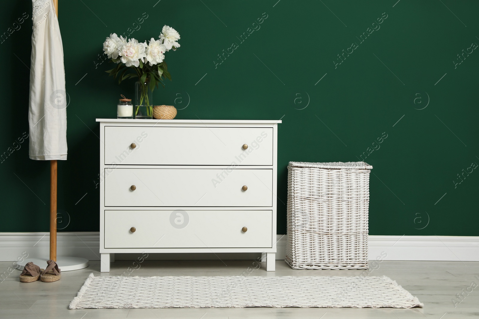 Photo of Modern white chest of drawers, coat stand and wicker basket near green wall indoors