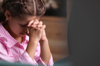 Photo of Cute little girl with hands clasped together praying indoors. Space for text