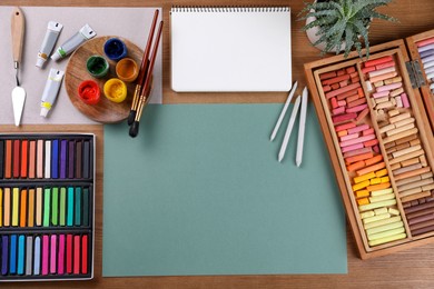 Blank sheet of paper, colorful chalk pastels and other drawing tools on wooden table, flat lay. Modern artist's workplace