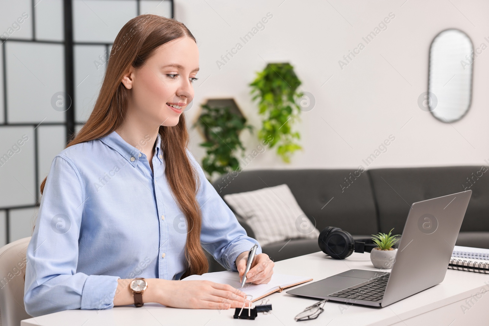 Photo of E-learning. Young woman taking notes during online lesson at white table indoors