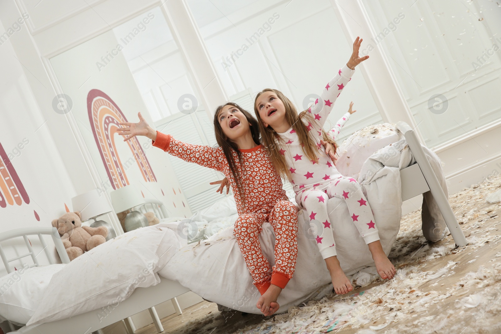 Photo of Cute little girls sitting on bed in messy room after pillow fight. Happy childhood