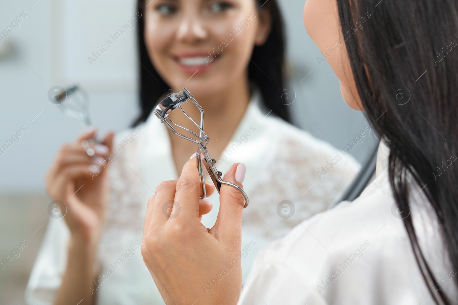 Photo of Beautiful young woman with eyelash curler near mirror indoors, closeup