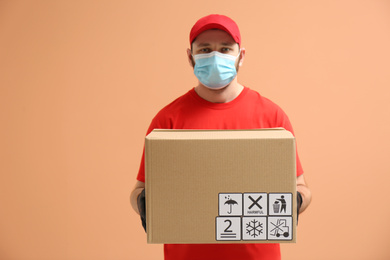 Photo of Courier in mask holding cardboard box with different packaging symbols on orange background. Parcel delivery