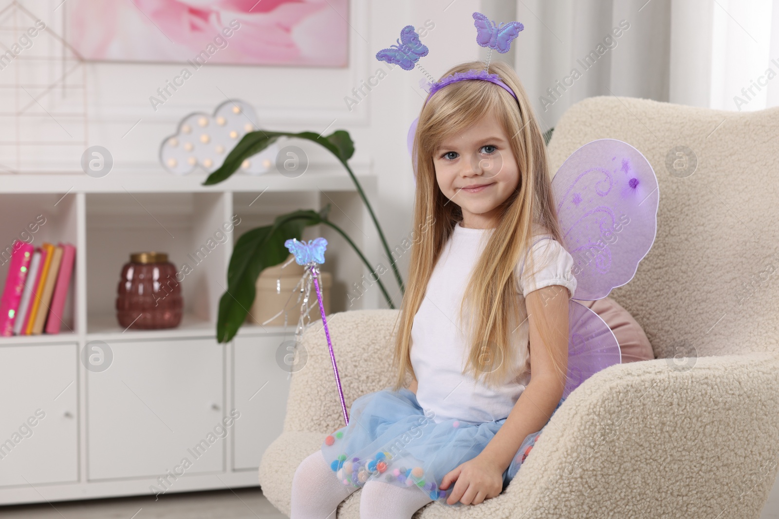 Photo of Cute little girl in fairy costume with violet wings and magic wand in armchair at home, space for text