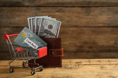 Photo of Credit card in shopping cart and wallet with dollar banknotes on wooden table, space for text. Cashback concept