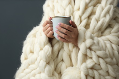 Woman covered with knitted plaid holding cup of hot drink on grey background, closeup