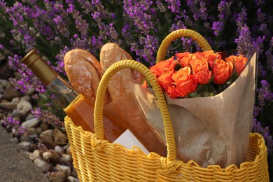 Yellow wicker bag with beautiful roses, bottle of wine and baguettes near lavender flowers outdoors, closeup