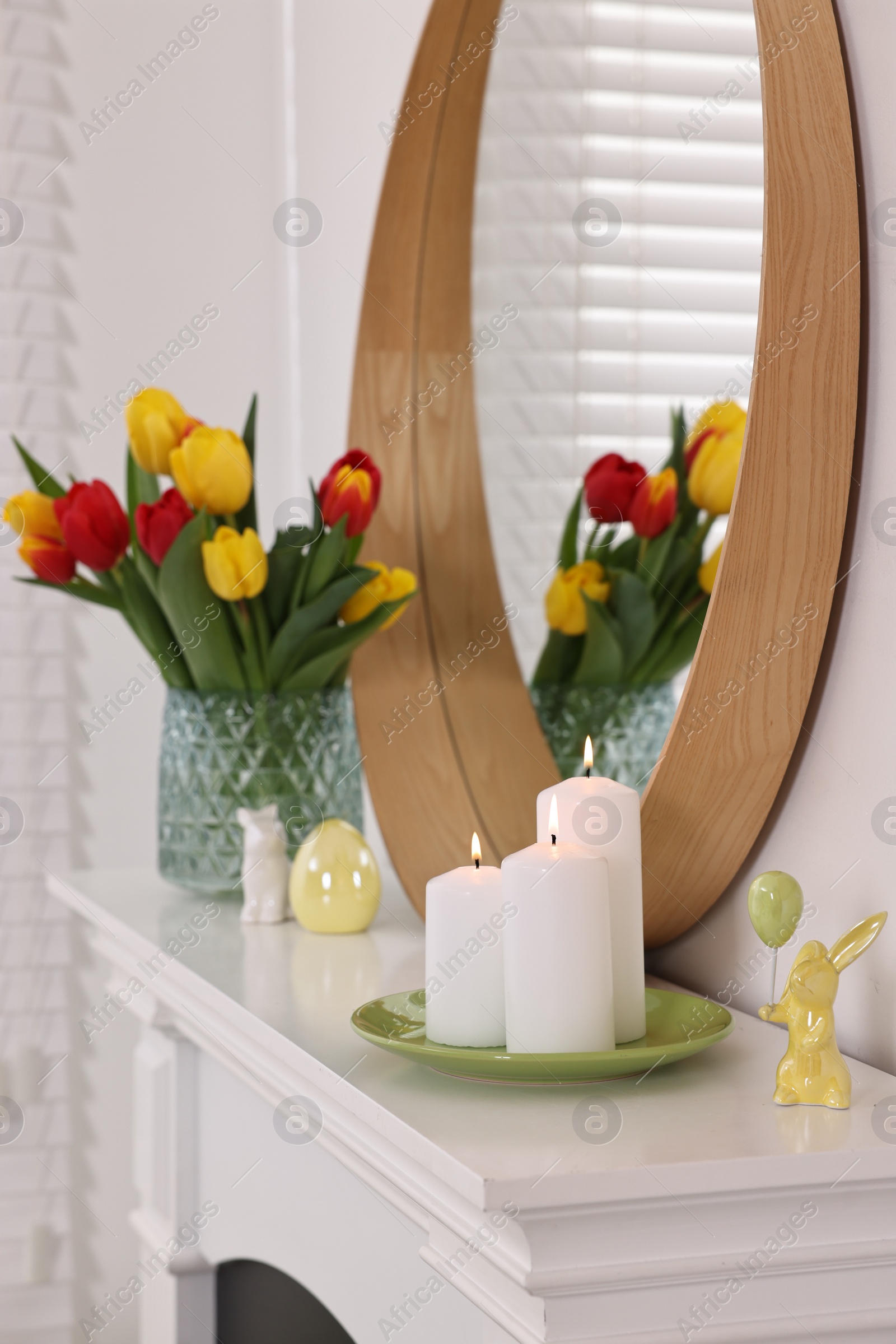 Photo of Easter decorations. Bouquet of tulips in vase, burning candles and figures on fireplace at home