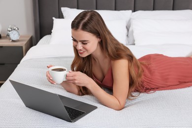 Photo of Happy woman with cup of coffee and laptop on bed in bedroom