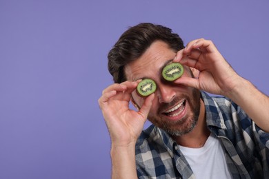 Photo of Man holding halves of kiwi near his eyes on purple background, space for text