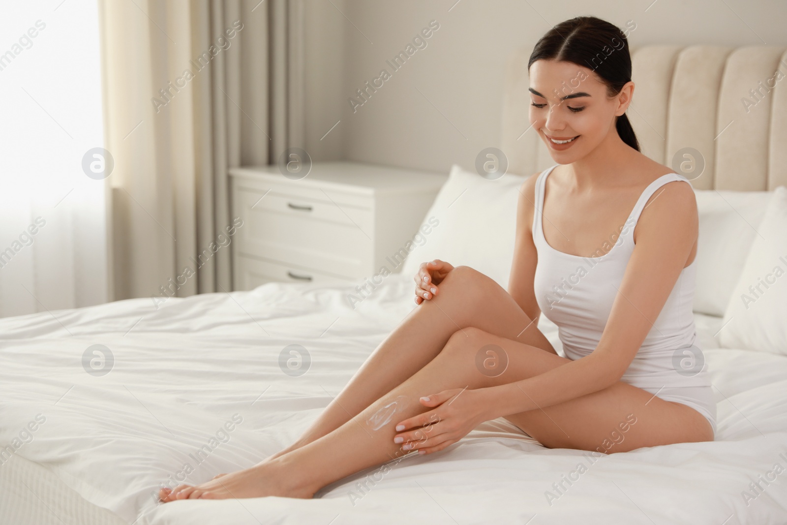 Photo of Young woman applying body cream onto her leg at home, space for text