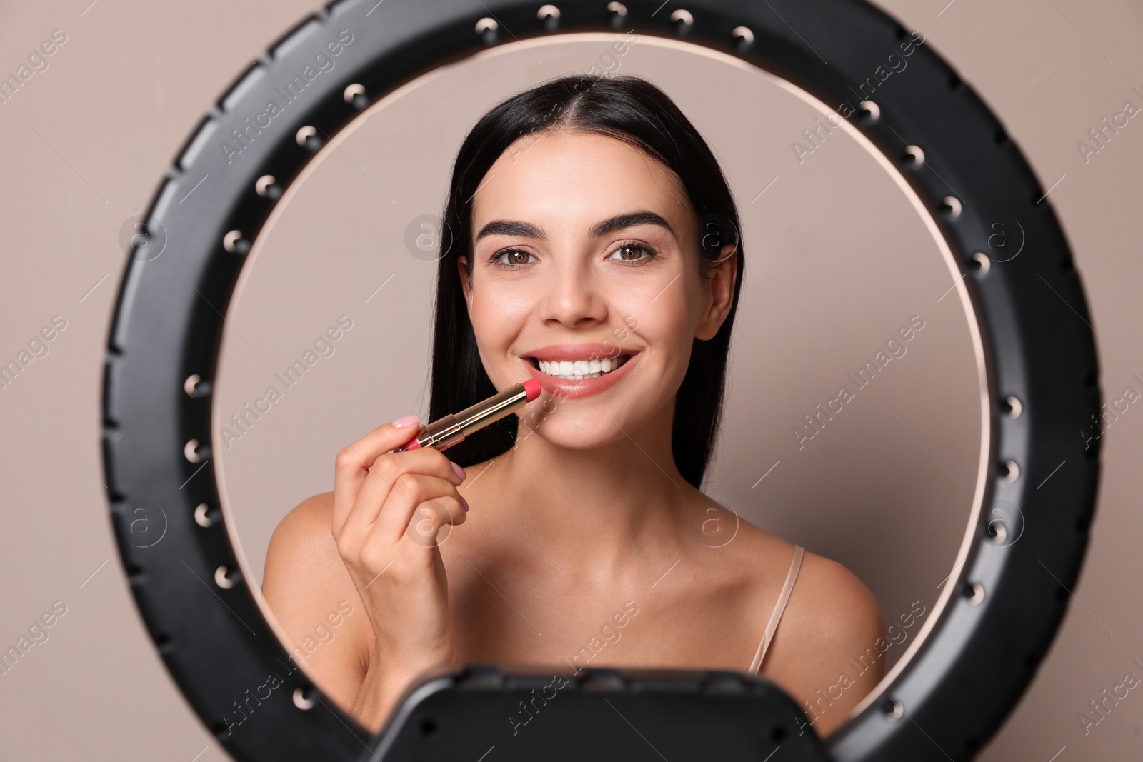 Photo of Beautiful young woman applying lipstick on beige background, view through ring lamp