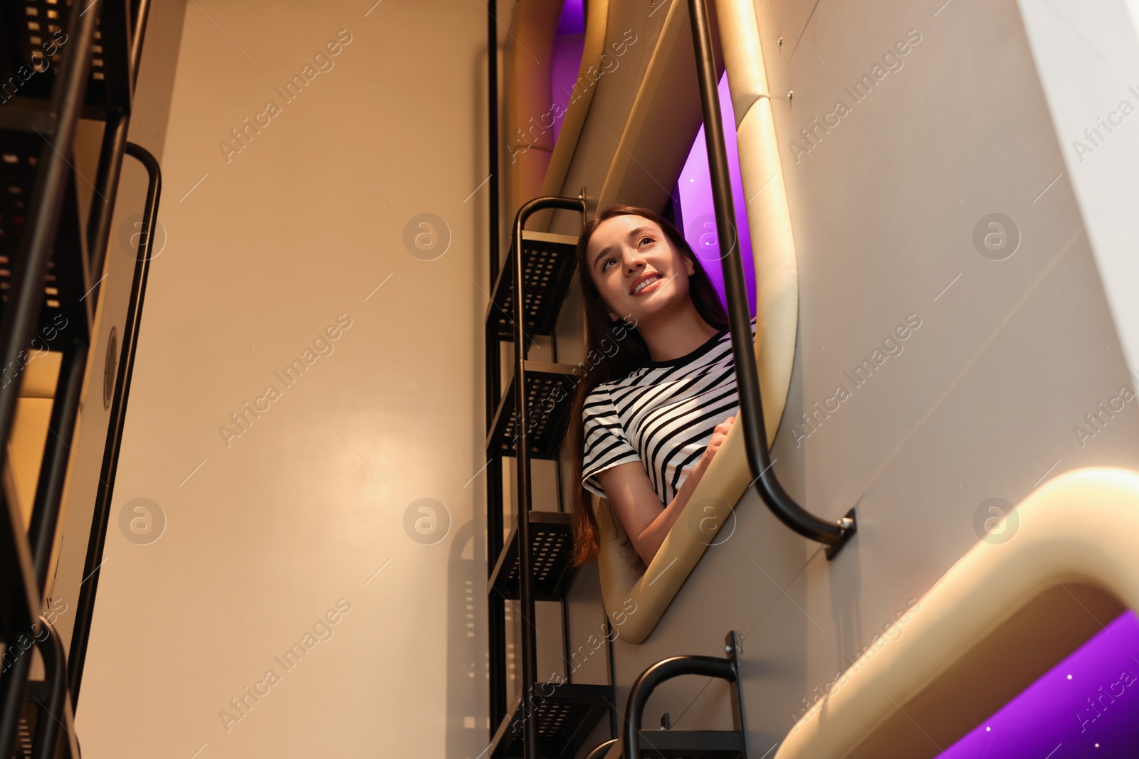 Photo of Happy young woman looking out of capsule in pod hostel, low angle view